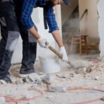 Close up shot of the legs of a man in a protective suit with a shovel in hand next to a bucket of debris left over from a house renovation