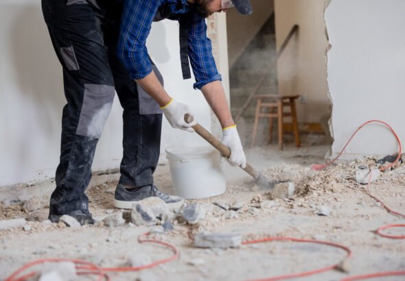 Close up shot of the legs of a man in a protective suit with a shovel in hand next to a bucket of debris left over from a house renovation