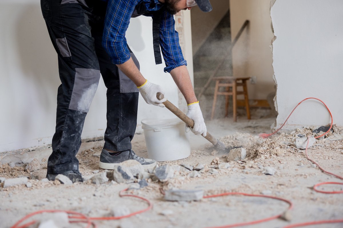 Close up shot of the legs of a man in a protective suit with a shovel in hand next to a bucket of debris left over from a house renovation